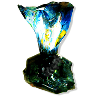 A unique, handcrafted fused glass lamp featuring vibrant shades of blue, green and white. The lamp has an abstract, sculptural shape with a flowing glass design that emits a soft, colourful glow